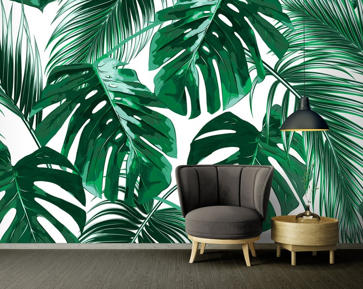 Fototapete mit großen grünen Monstera- und Palmenblättern in einem modern eingerichteten Raum mit grauem Sessel und Holzbeistelltisch.