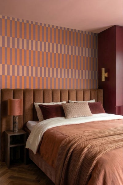 Modernes Schlafzimmer mit gepolstertem Bett mit Samtkopfteil, geometrisch gemusterter Tapete, Nachttisch mit Lampe und Einbauschrank.