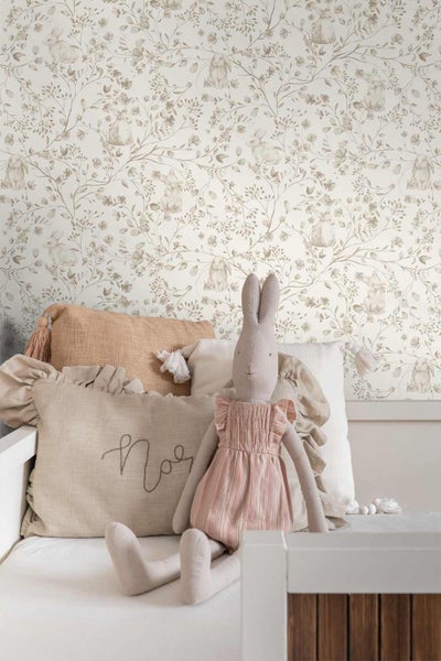 Vliestapete mit Hasenmotiven und floralem Muster in einem hellen Kinderzimmer hinter einem Kinderbett mit Kissen und Stoffhase.