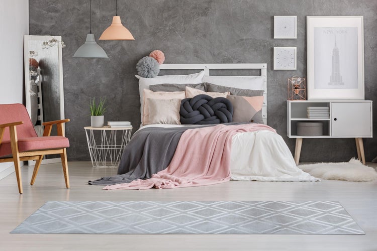 Modernes Schlafzimmer mit grau strukturierter Wand, weißem Bettgestell, rosa Sessel und einem Teppich mit Rautenmuster.