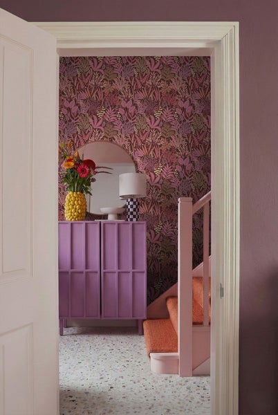 Ein Flur mit einem lilafarbenen Schrank, einem Spiegel, einer Vase mit Blumen und einer Tapete mit Blattmuster.