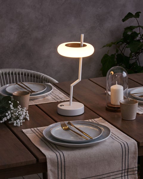 Moderne LED Tischleuchte in Weiß mit ringförmigem Leuchtkopf auf einem gedeckten Holztisch mit Geschirr und Dekoration.