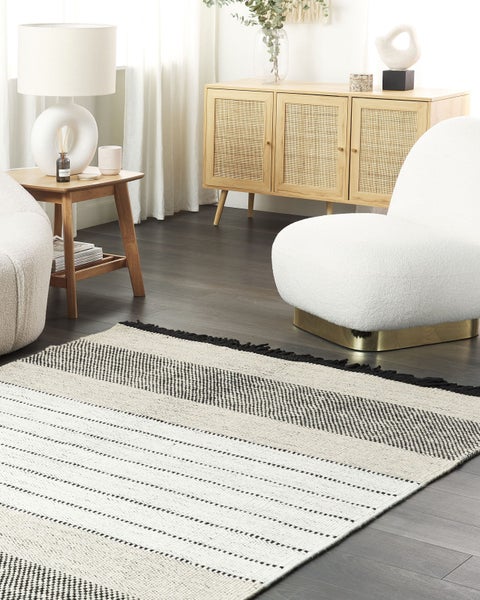 Gewebter Teppich mit schwarz-weißen Streifen in hellem Wohnzimmer mit weißem Bouclé-Sessel, Holz-Sideboard und Beistelltisch.
