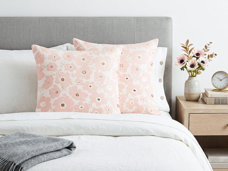 Zwei quadratische Dekokissen mit Blumenmuster auf einem gemachten Bett mit grauem Kopfteil und Nachttisch.