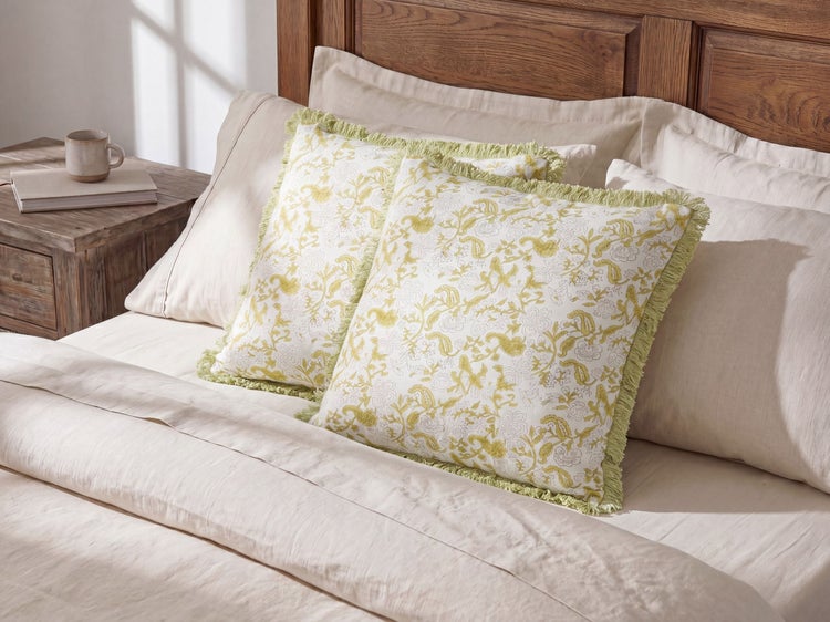 Zwei quadratische Dekokissen mit floralem Muster und Fransenrand in einem hellen Schlafzimmer auf einem Bett mit hölzernem Kopfteil.