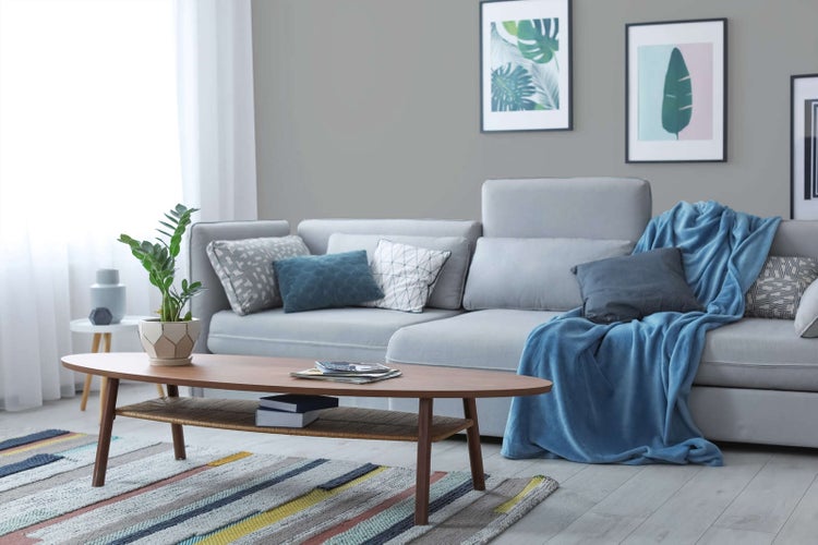 Modernes Wohnzimmer mit grauem Ecksofa, ovalem Couchtisch aus Holz, gemustertem Teppich und botanischen Bildern an der grauen Wand.