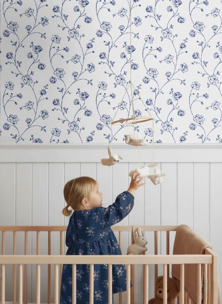 Kinderzimmer mit floraler Tapete, weißen Holzpaneelen und Holzbabybett. Ein Kind greift nach einem Mobile mit Stofftieren.