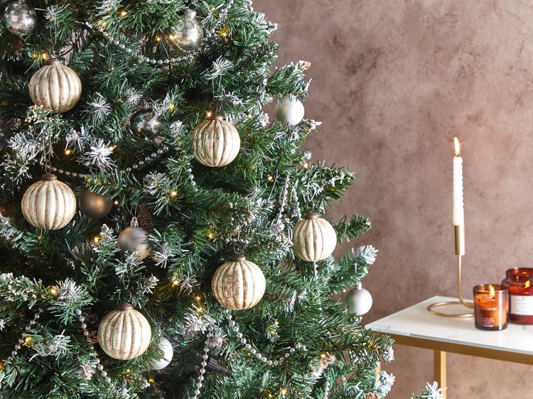 Grüner künstlicher Weihnachtsbaum mit gerippten Kugeln und silbernen Perlenketten neben einem Beistelltisch mit Kerzen.