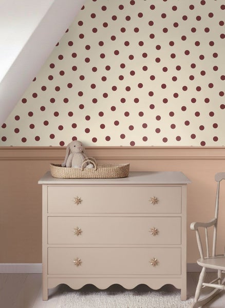 Hell eingerichtetes Kinderzimmer mit gepunkteter Tapete an einer Dachschräge, einer Kommode mit Sternknöpfen und einem Stoffhasen in einem Körbchen.