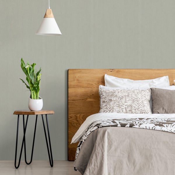 Helles Schlafzimmer mit strukturierter hellgrüner Vliestapete. Zu sehen sind ein Bett mit Holz-Kopfteil, ein Beistelltisch mit Pflanze und eine Lampe.