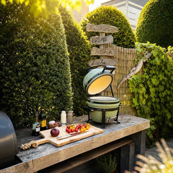 Offener grüner Keramik-Kamado-Grill auf rustikaler Außenküche aus Holz mit Gemüse, Öl und dekorativem Schild im Garten.