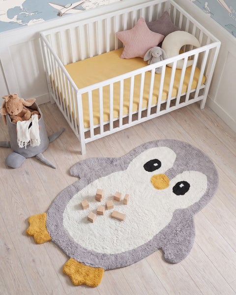 Kinderzimmer mit weißem Gitterbett, Stoffkörbchen mit Kuscheltieren und einem Pinguin geformten Teppich mit Holzbausteinen auf hellem Boden.