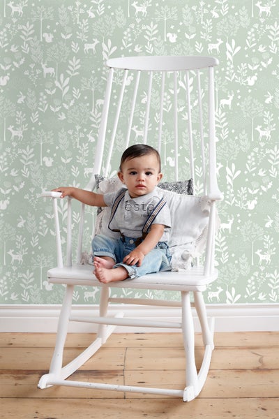 Baby sitzt in weißem Schaukelstuhl vor Wandtapete mit Waldmotiven