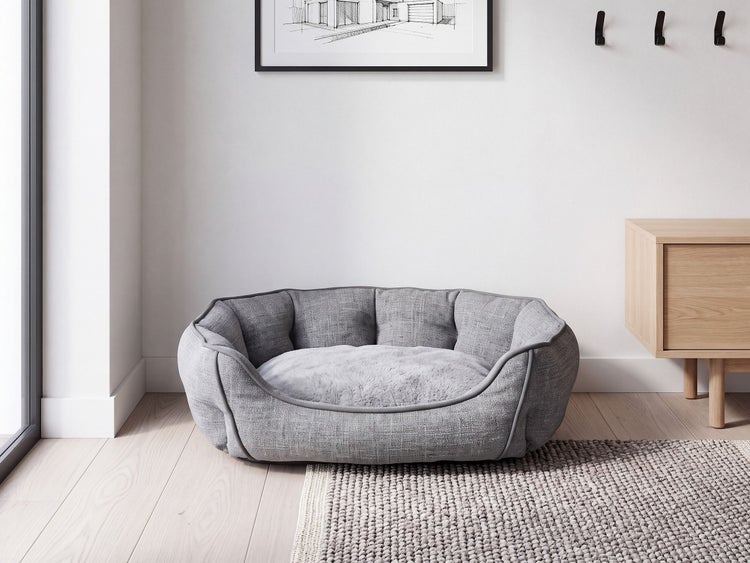 Ein weiches Hundebett mit erhöhten Rändern in einem modernen Wohnraum mit Holzboden, neben einem Hängeschrank und einem Wandbild.
