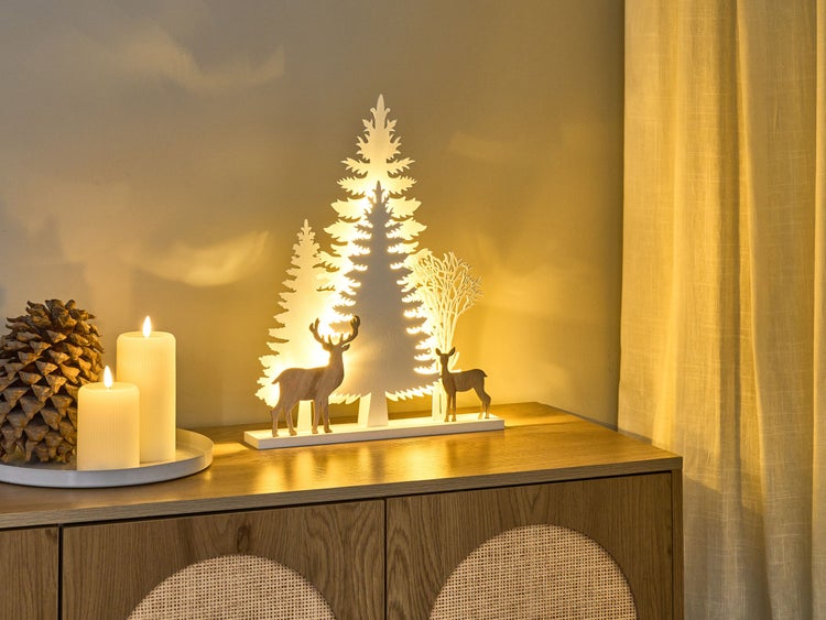 Weihnachtsdekoration mit Tannenbaum, Hirschfiguren und Kerzen auf einem Holzschrank.