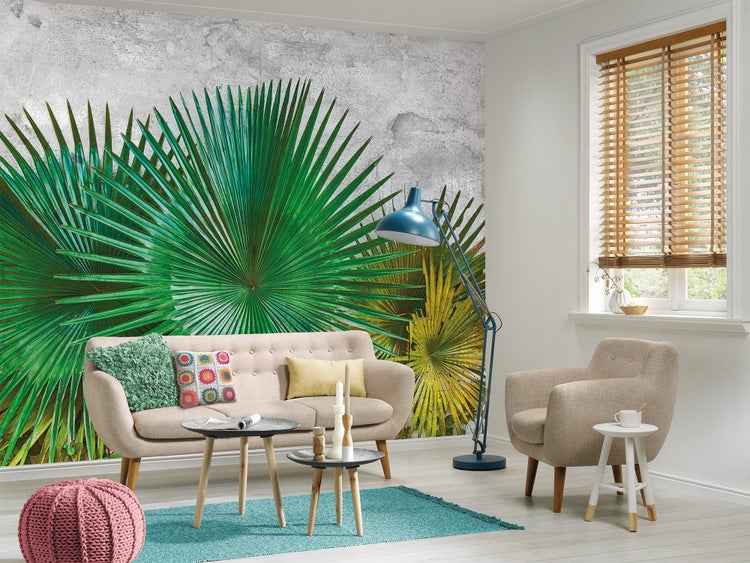 Modernes Wohnzimmer mit einer Fototapete mit großen grünen Palmblättern auf grauem strukturiertem Hintergrund, beigem Sofa und blauer Stehlampe.
