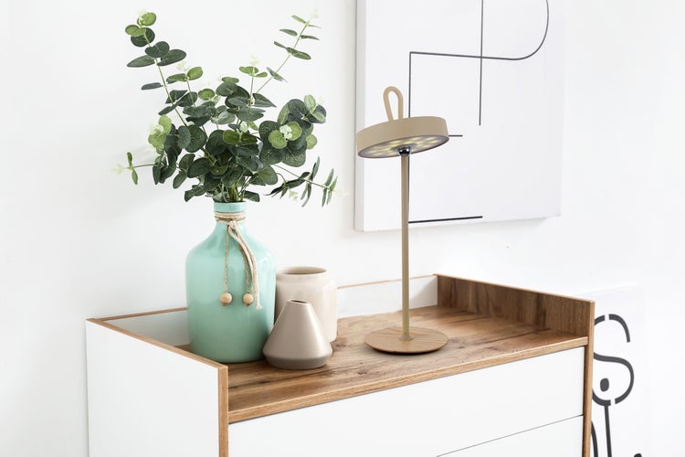 Dekoratives Arrangement mit Tischleuchte, Vase und Kunstpflanzen auf einer Kommode.