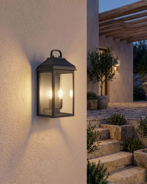 Klassische Außenwandleuchte in Laternenform aus Metall mit Glaseinsätzen, montiert an einer hellen Hauswand auf einer Terrasse.