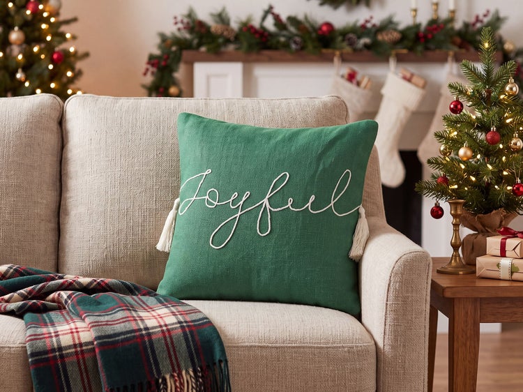 Weihnachtskissen mit Schriftzug Joyful und Quasten auf einem Sofa in einem weihnachtlich dekorierten Wohnzimmer.