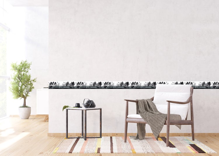 Selbstklebende Wandbordüre mit Wald-Silhouette und Elchen an einer Wand in einem hellen Zimmer mit Sessel und Beistelltisch mit Teekanne.