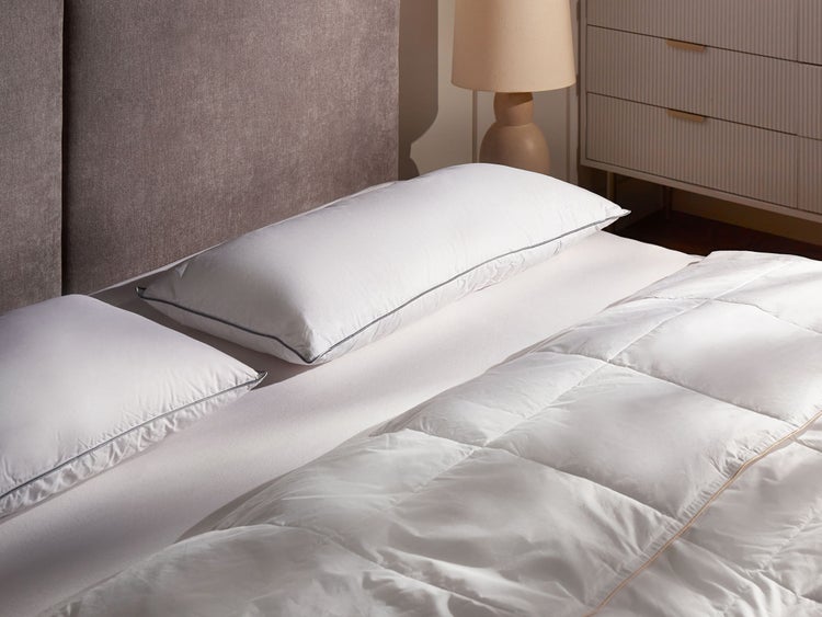 Bett mit zwei weißen Kopfkissen mit dunkler Paspelierung, einer gesteppten weißen Decke und einem gepolsterten Kopfteil.