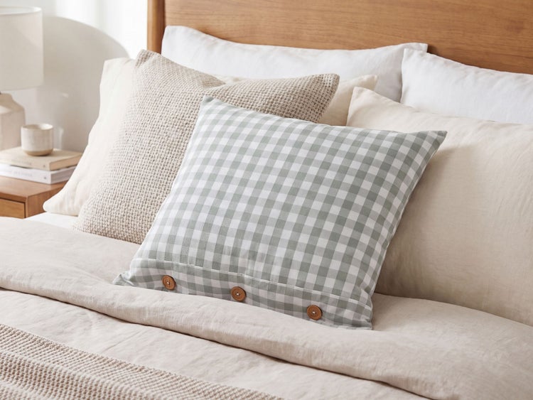 Quadratisches Zierkissen mit Vichy Karo Muster und Knopfleiste auf einem bezogenen Bett mit weiteren Kissen und hölzernem Kopfteil.