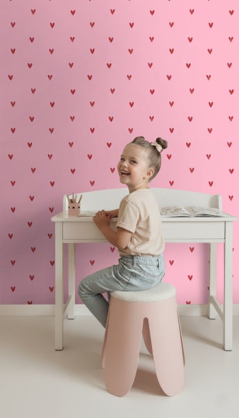 Mädchen sitzt an einem Schreibtisch vor einer Wandtapete mit Herzmotiven
