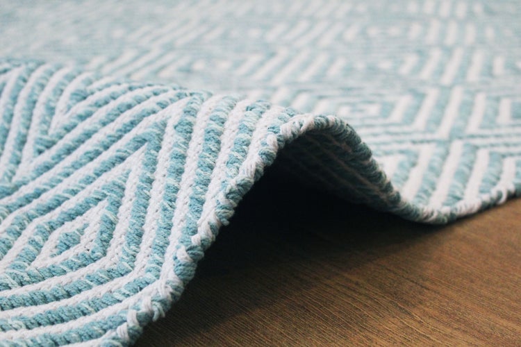 Nahaufnahme eines gewebten Teppichs mit geometrischem Muster in Hellblau und Weiß, gefaltete Kante auf Holzfußboden.