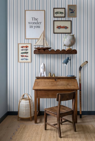 Kinderzimmer mit blau-weiß gestreifter Tapete, einem Holzsekretär mit Stuhl, Wandbildern und dekorativen Objekten wie Segelboot und Globus.