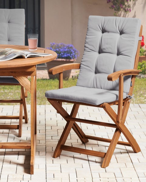 Gartenklappstuhl aus Holz mit grauer Polsterauflage auf einer gepflasterten Terrasse neben einem runden Holztisch.