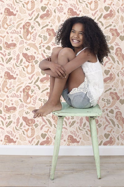 Mädchen sitzt vor einer Tapete mit Meerjungfrauenmotiven auf einem Hocker