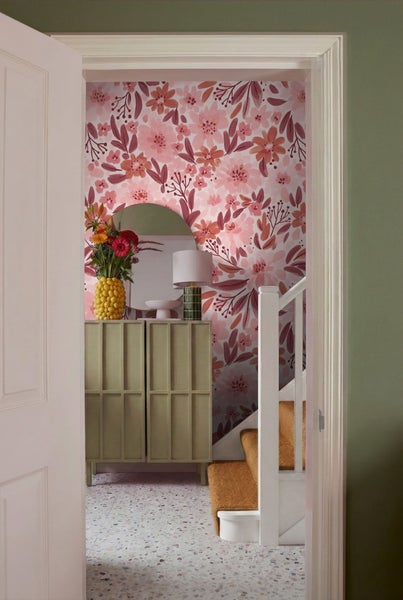 Blick durch eine geöffnete Tür auf eine dekorierte Diele mit Blumentapete, Kommode und Treppe