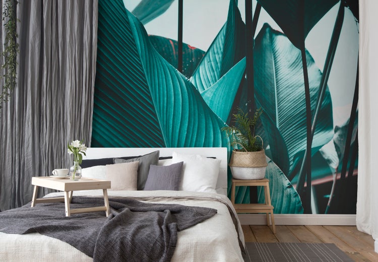 Fototapete mit großem tropischem Blattmotiv in einem Schlafzimmer mit weißem Bett, grauen Textilien und Holzakzenten.