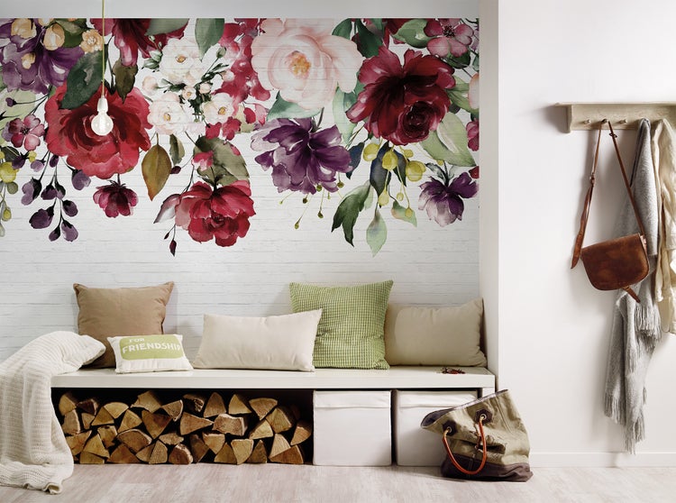 Vlies-Fototapete mit Aquarell-Blumenmuster auf weißer Ziegelwand über einer Sitzbank mit Brennholz und Kissen in einem hellen Raum.