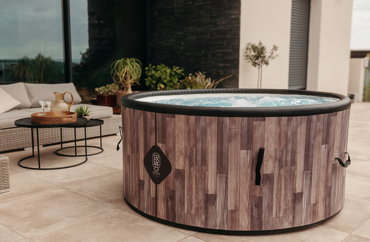 Runder aufblasbarer Whirlpool in Holzoptik auf einer modernen Terrasse mit Loungemöbeln. Coco Spa Logo.