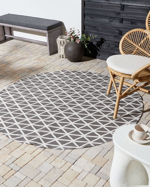 Runder Outdoor-Teppich mit Dreiecksmuster auf einer gepflasterten Terrasse mit Gartenbank, Rattansessel und Beistelltisch.
