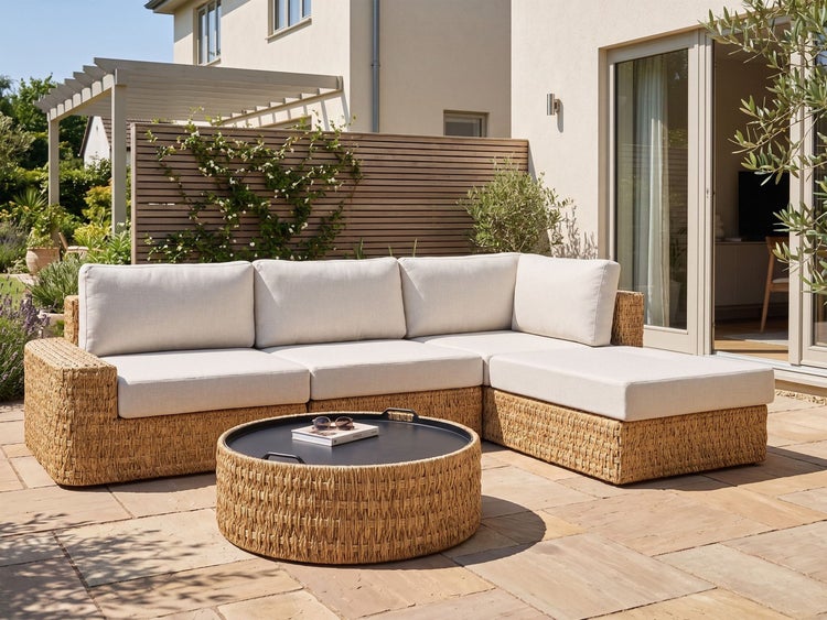 Eck-Loungegruppe aus Geflecht mit hellen Kissen und rundem Couchtisch auf einer gepflasterten Terrasse vor einer Holzwand.