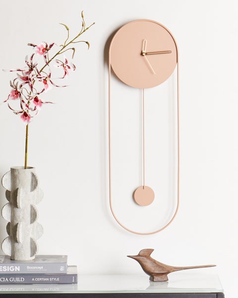 Moderne Wanduhr mit Pendel und schmalem Metallrahmen, neben einer strukturierten Vase mit Orchidee und einer Vogelfigur aus Holz.