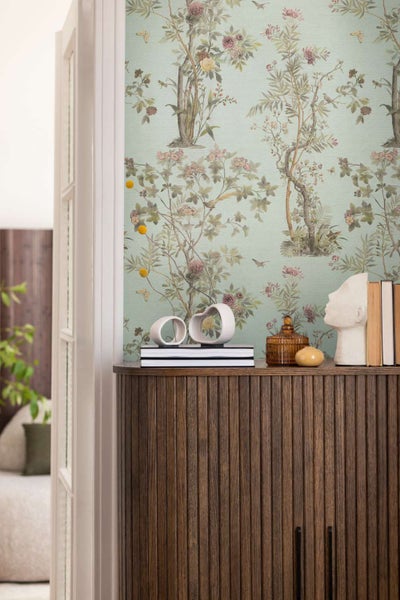 Wohnraum mit floraler Vliestapete im botanischen Stil und einer Kommode aus geriffeltem Holz, dekoriert mit Skulpturen und Büchern.