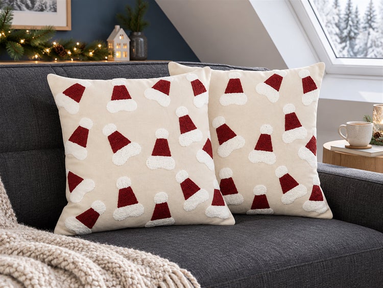 Zwei quadratische Dekokissen mit Weihnachtsmützenmotiv auf einem Sofa vor weihnachtlicher Dekoration.