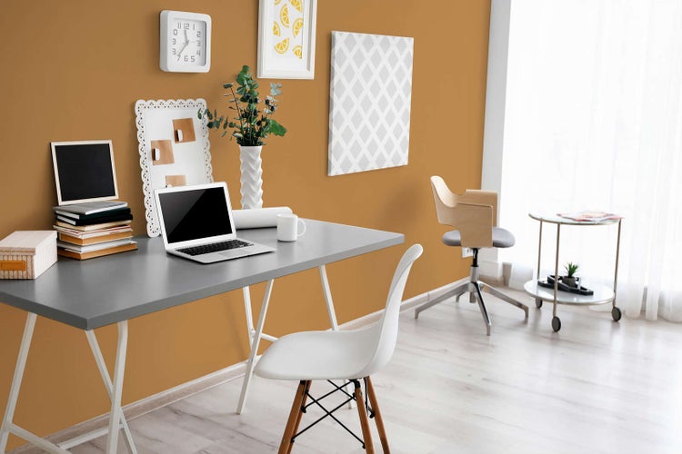 Modernes Arbeitszimmer mit ockerfarbener Wand, grauem Schreibtisch, weißem Stuhl und hellem Boden. Organisierter Arbeitsplatz mit viel Tageslicht.