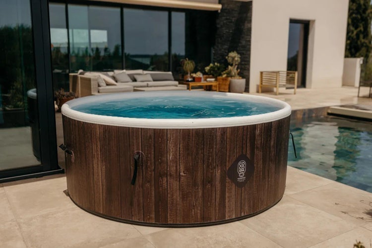 Runder Whirlpool in Holzoptik auf einer modernen Terrasse mit hellen Fliesen neben einem Pool und Gartenmöbeln.