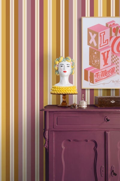 Gestreifte Tapete in Ocker, Rosa und Rot an einer Wand hinter einer lila Kommode mit dekorativer Büste und gerahmtem Bild.