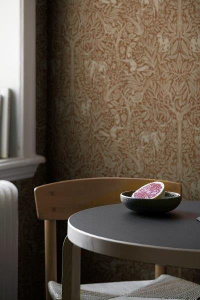 Ein Schlafzimmer mit einem Bett mit einem Korbstrohkopfende, einem Nachttisch mit einer Lampe, einer Uhr, einer Figur und Topfpflanzen sowie einer gemusterten Tapete.