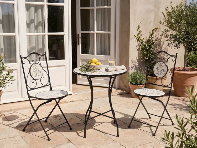 Bistro-Set aus rundem Tisch und zwei Klappstühlen, schwarzes Metallgestell mit Mosaikmuster, auf einer hellen Steinterrasse mit Pflanzen.