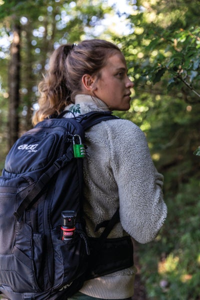Eine Person mit einem schwarzen Rucksack, der mit einem Zahlenschloss gesichert ist und ein Abwehrspray trägt.