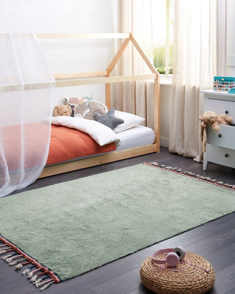 Hellgrüner Kinderteppich mit bunten Fransen in einem hellen Kinderzimmer mit hölzernem Hausbett, weißem Baldachin und hellen Vorhängen.