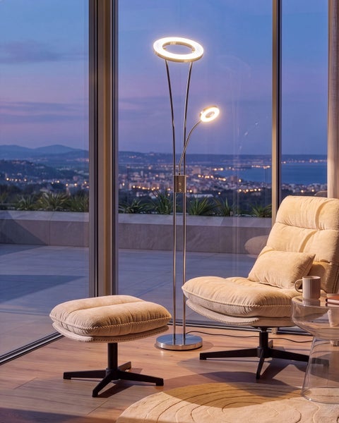 Moderne LED Deckenfluter Stehlampe mit Leseleuchte neben einem bequemen Sessel mit Fußhocker vor einem großen Fenster mit nächtlicher Aussicht
