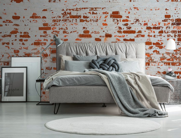 Modernes Schlafzimmer mit grauem Polsterbett, verschiedenen Textilien, rundem Teppich und einer Wand in rustikaler Backsteinoptik.
