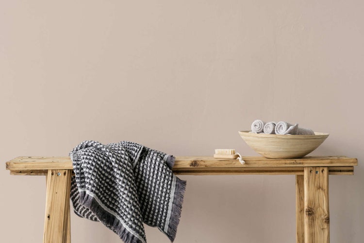 Rustikale Holzbank mit einem gemusterten Waffelhandtuch, einer Holzschale mit Gästehandtüchern und einer Nagelbürste vor einer schlichten Wand.